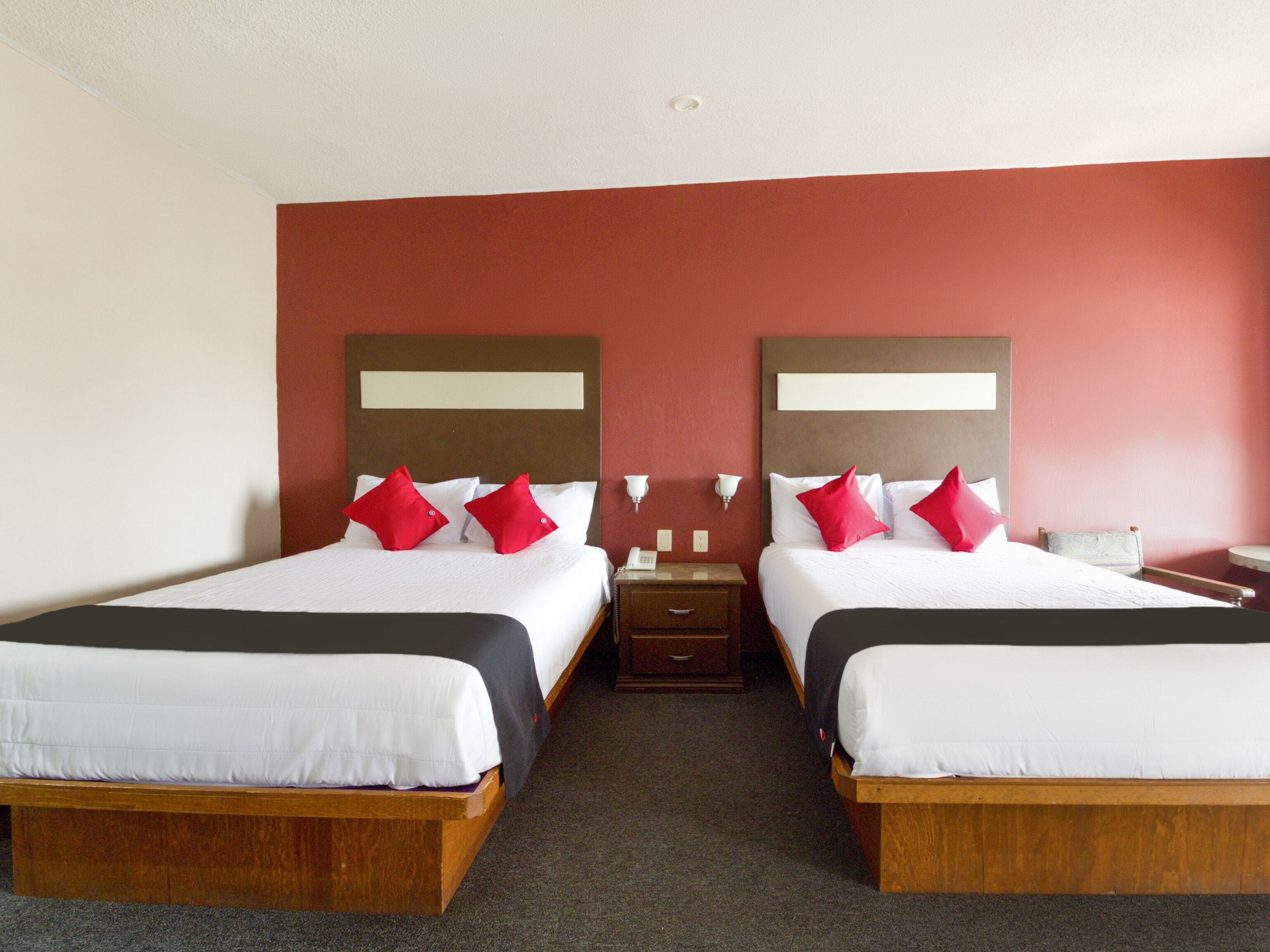 A hotel room with two beds, each with white bedding and red pillows, and a wooden headboard.