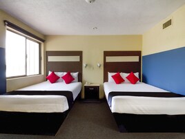 Habitación de hotel con dos camas, cada una con ropa de cama blanca y almohadas rojas, una mesita de noche con lámpara y una ventana con persianas.