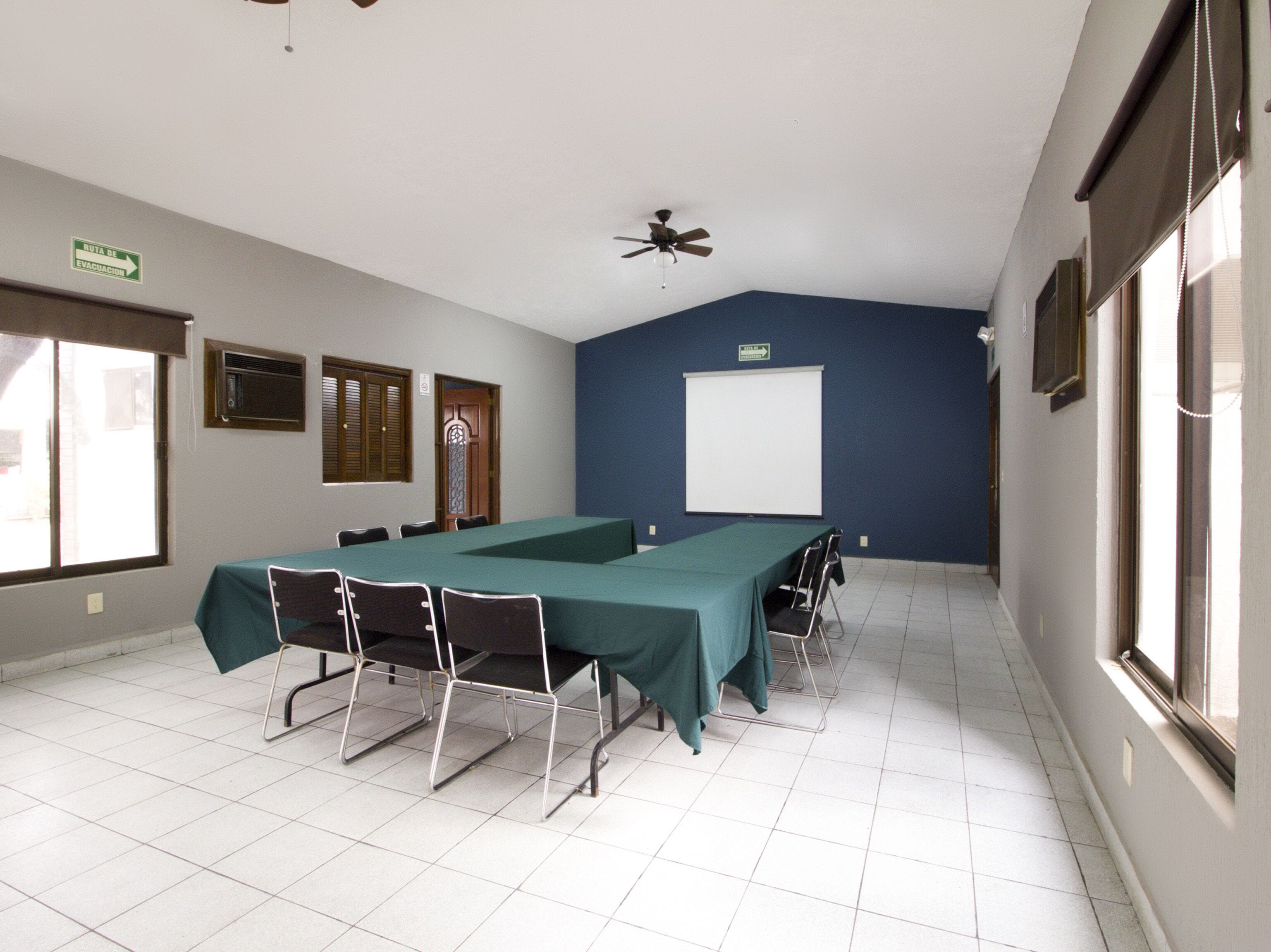 Un salón de conferencias con una larga mesa, sillas, una pantalla de proyector y un ventilador de techo.