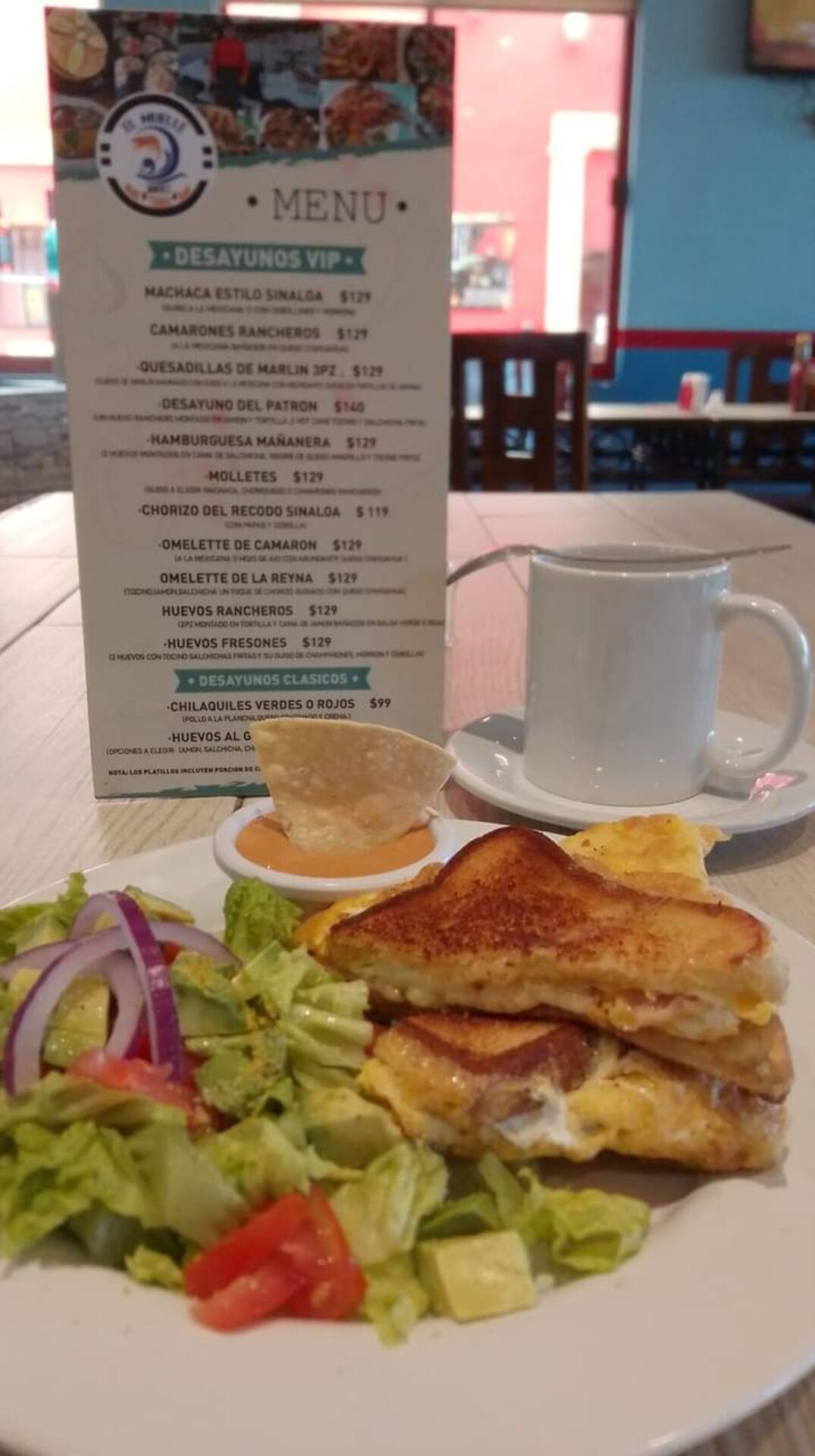 Un menú con diferentes opciones de comida y sus precios, un plato con un sándwich a la plancha, una ensalada y una taza de café.