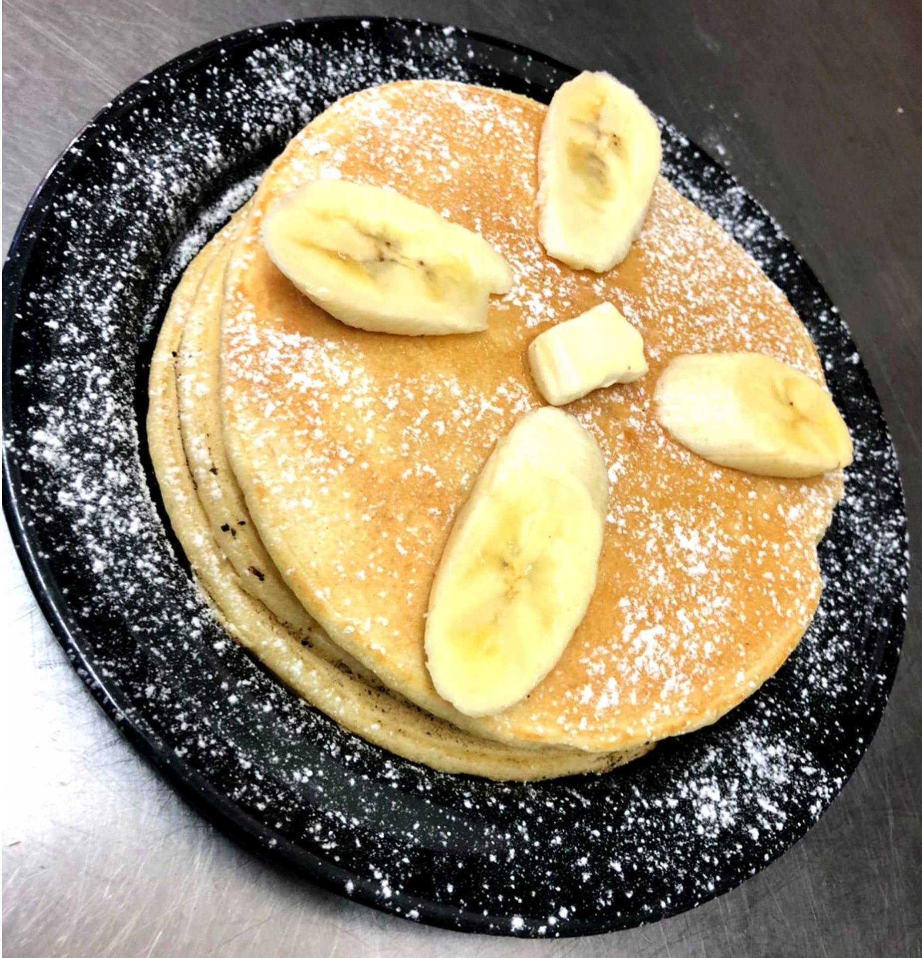 En hög med pannkakor, toppad med bananskivor och pudersocker, serverad på en svart tallrik.