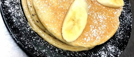 En hög med pannkakor, toppad med bananskivor och pudersocker, serverad på en svart tallrik.