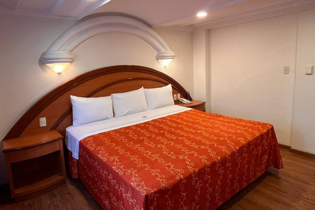 A bedroom with a wooden headboard, a red patterned bedspread, two white pillows, a bedside table with a lamp, and a nightstand with a phone.