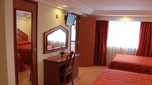 A hotel room with a wooden desk, two beds with red bedding, a large mirror, and a television on the wall.