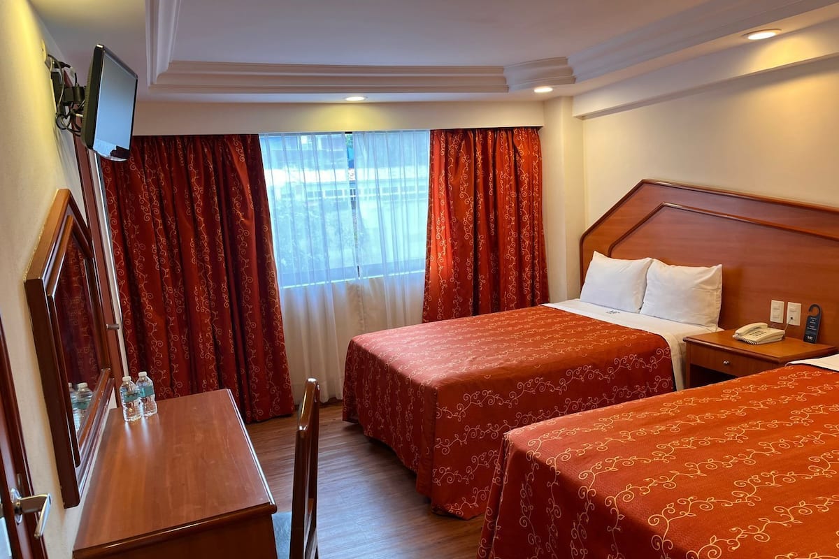A hotel room with two beds, a desk, a chair, a TV mounted on the wall, and a window with red curtains.
