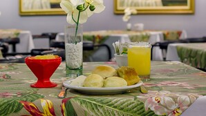 A dining table with a floral tablecloth, a plate of food, a red bowl, a glass of orange juice, and a vase with white flowers. - Hotel Nacional Inn Curitiba Torres (Curitiba)