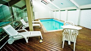 An indoor swimming pool with wooden decking, white lounge chairs, and a table. - Hotel Nacional Inn Curitiba Torres (Curitiba)