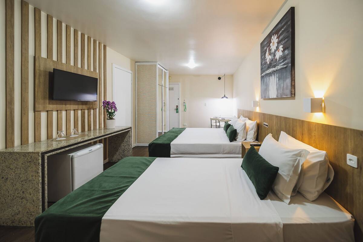Ein Hotelzimmer mit zwei Betten, einem an einer Holzplatte montierten Fernseher, einem kleinen Schreibtisch mit Kühlschrank und einem Bild an der Wand.