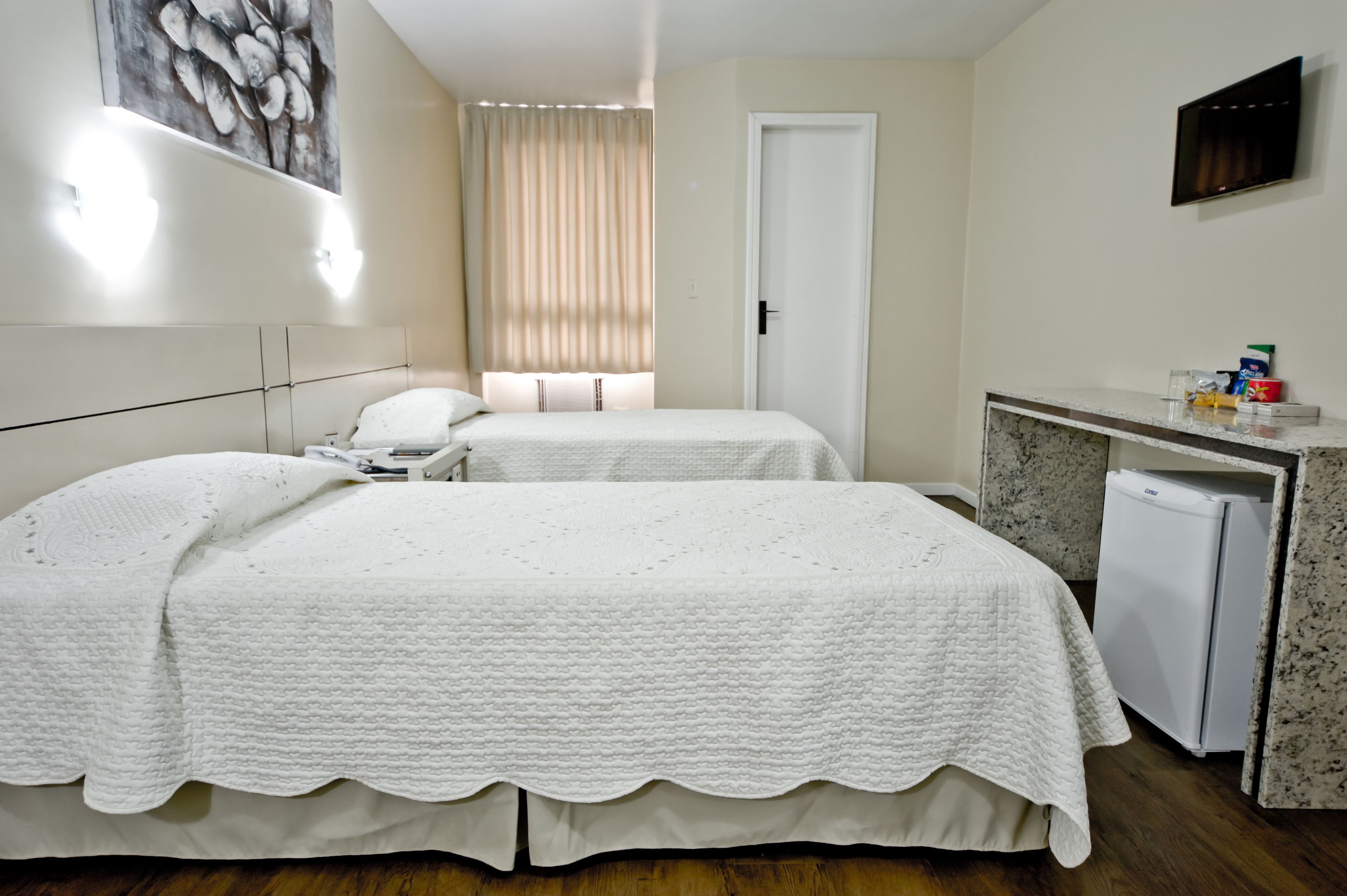 A hotel room with two beds, a small kitchenette, a TV, and a painting on the wall.