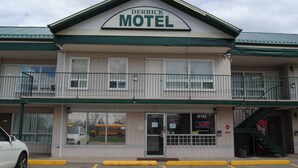 Un motel doté d’un toit végétalisé et d’une enseigne indiquant « Derrick Motel ». Un parking se trouve devant, et une voiture est garée sur le côté gauche.