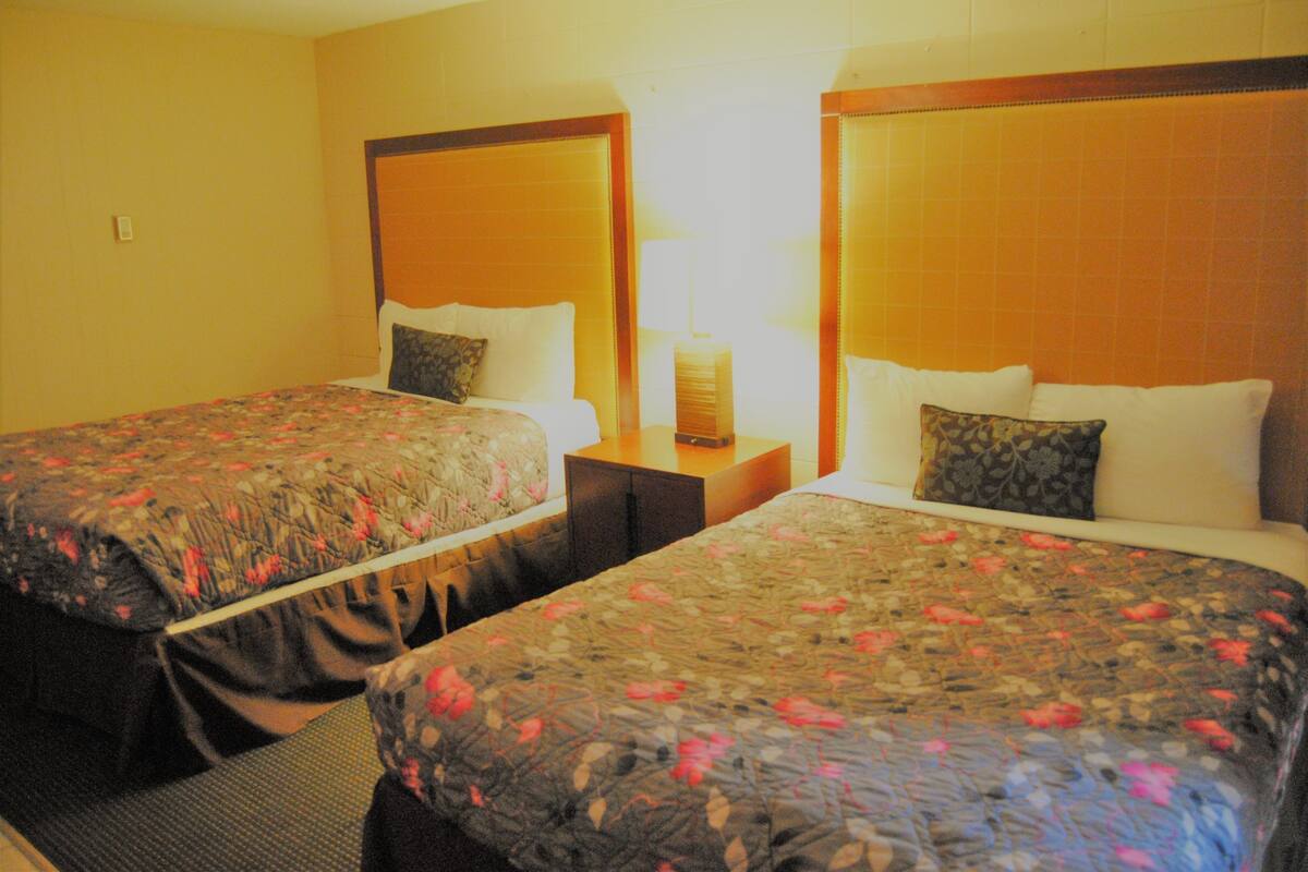 Dois quartos de hotel lado a lado, com colchas estampadas com flores, cabeceiras de madeira e uma mesa de cabeceira entre eles.