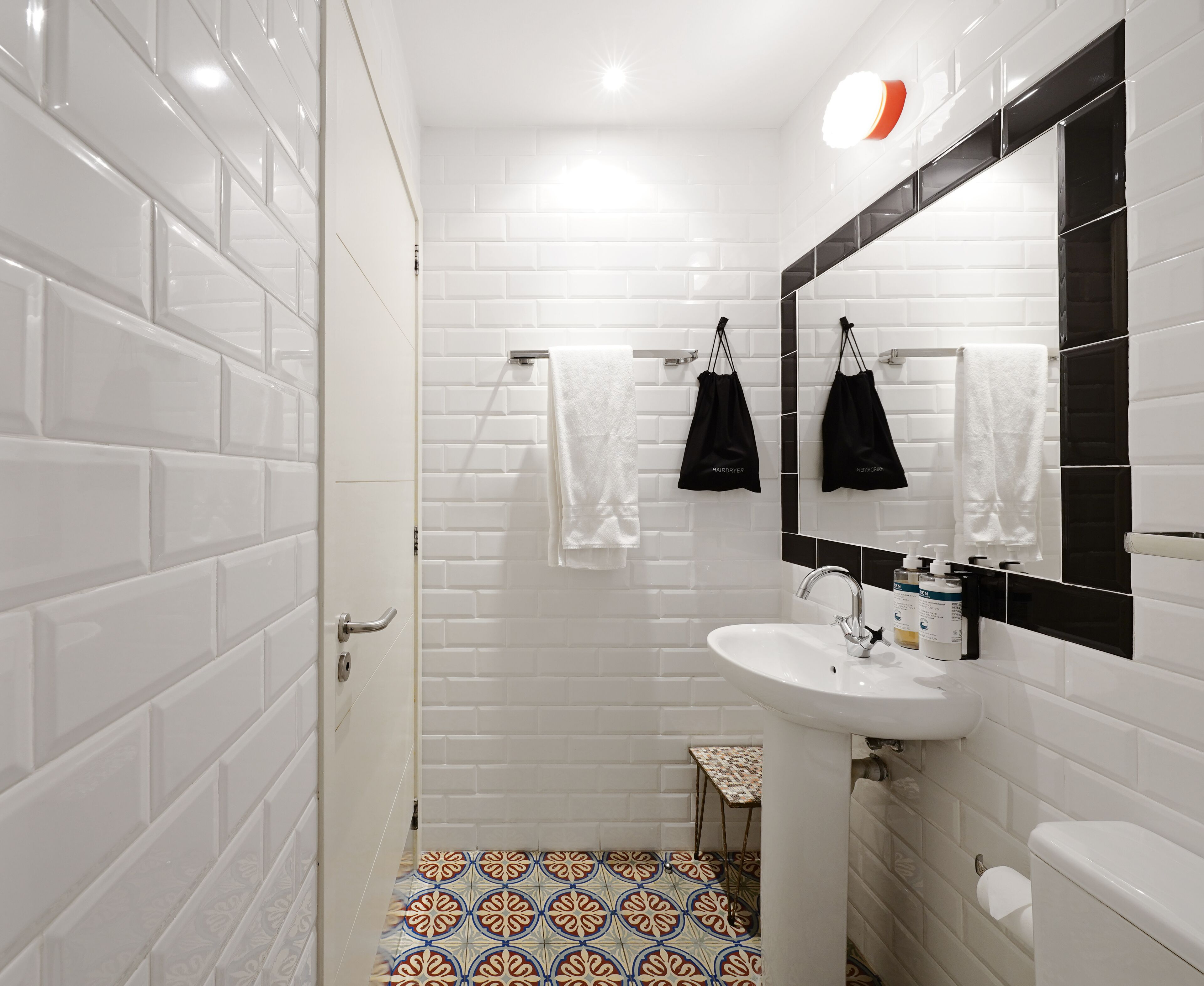Un baño con azulejos blancos tipo metro, un lavamanos blanco, un espejo y una pequeña mesa con mantel estampado.