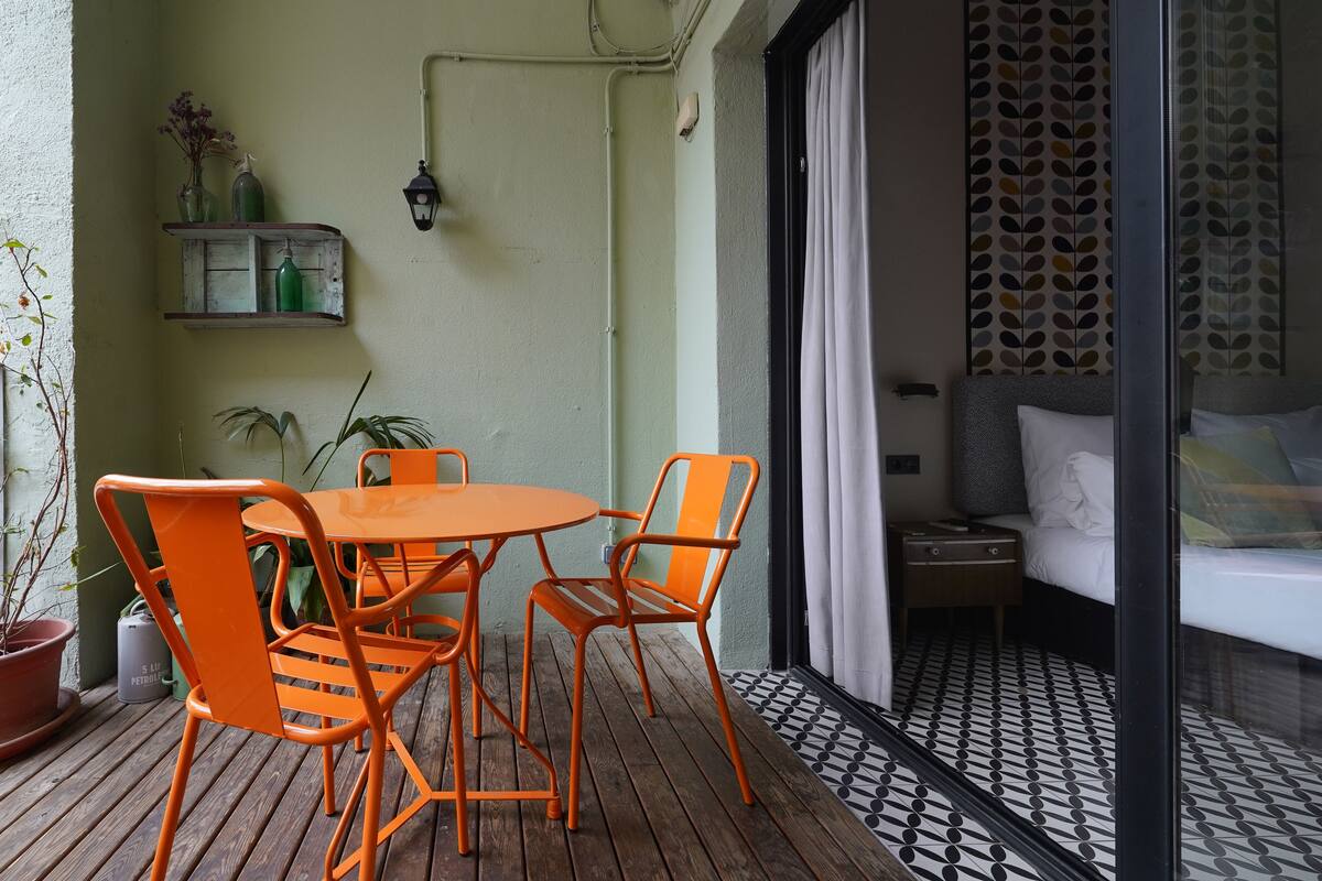 Una habitación pequeña y acogedora con una mesa de madera y sillas color naranja, una cama con colchón blanco y un suelo con estampado.