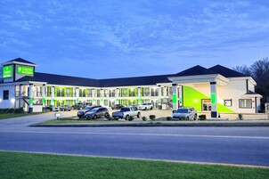 Un hotel de varios pisos con un diseño moderno, que cuenta con un estacionamiento con varios autos y una entrada iluminada con luces verdes.