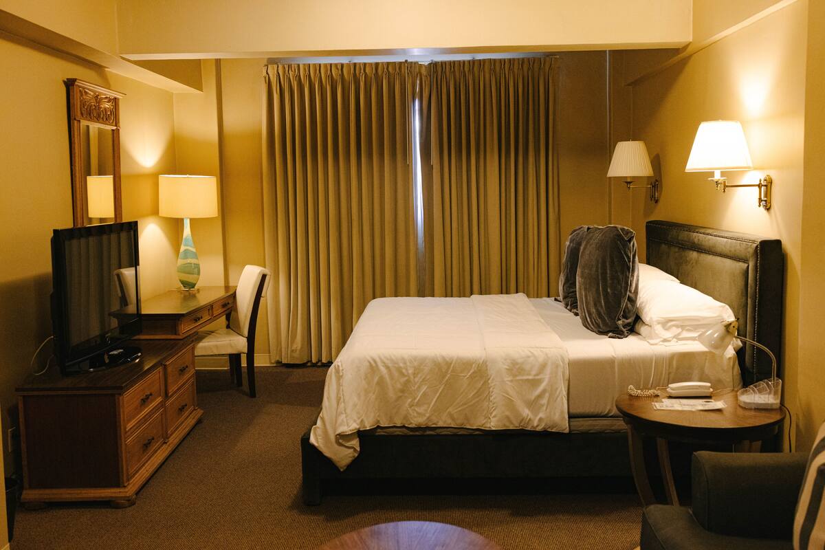 Una habitación de hotel con cama, escritorio con lámpara, silla, televisor y ventana con cortinas.