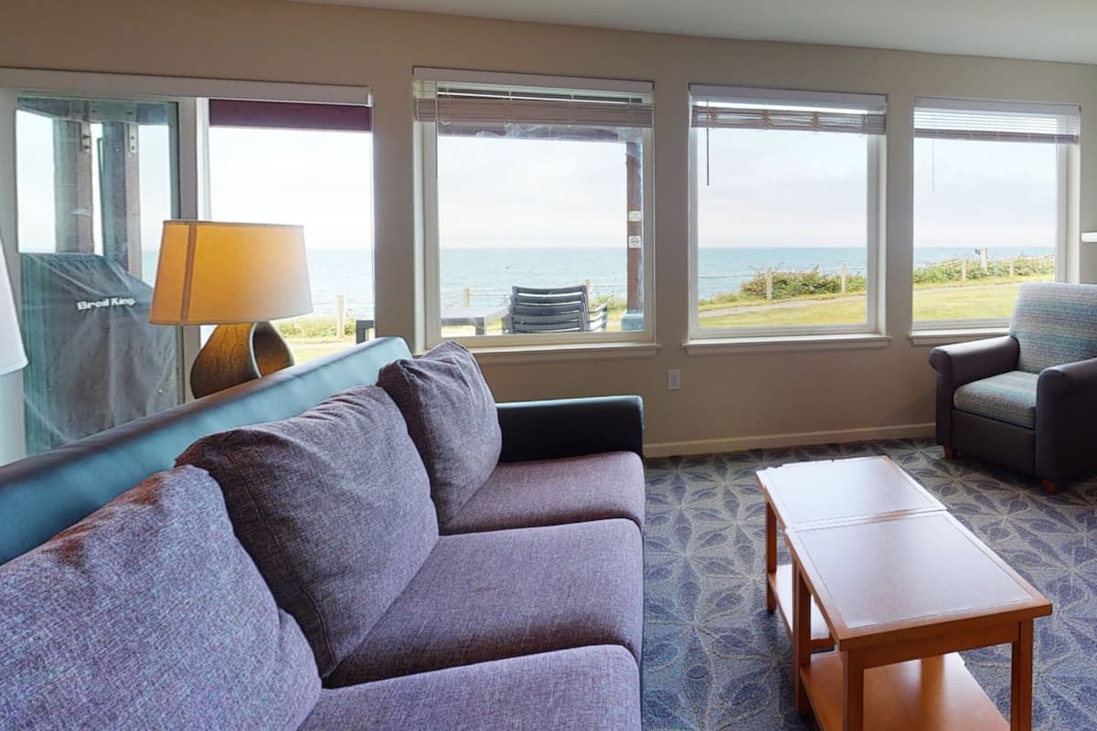 A room with a sofa, armchair, and a table. There are large windows with a view of the sea.