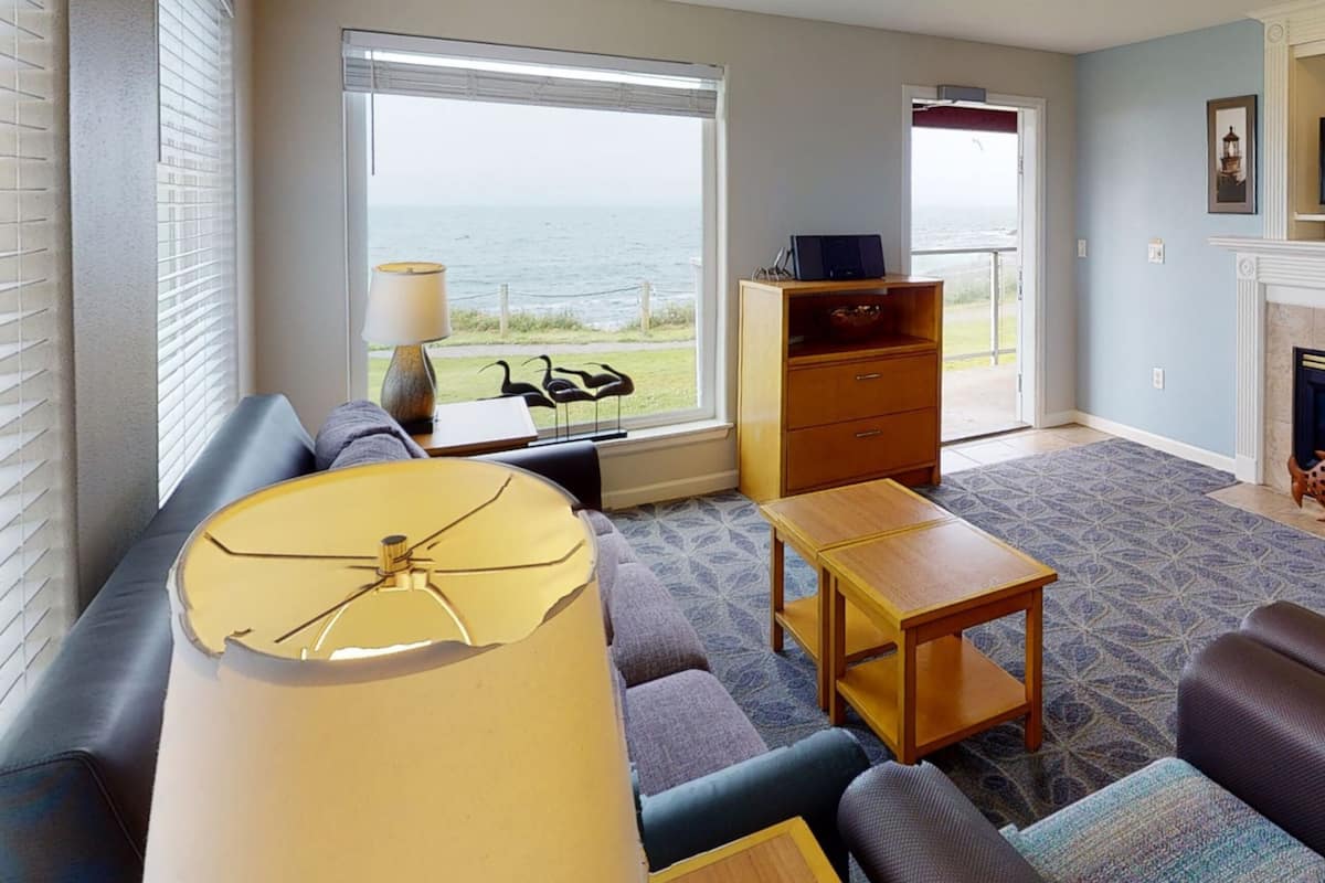 A living room with a sofa, a coffee table, a lamp, a wooden cabinet, and a view of the ocean through a large window.