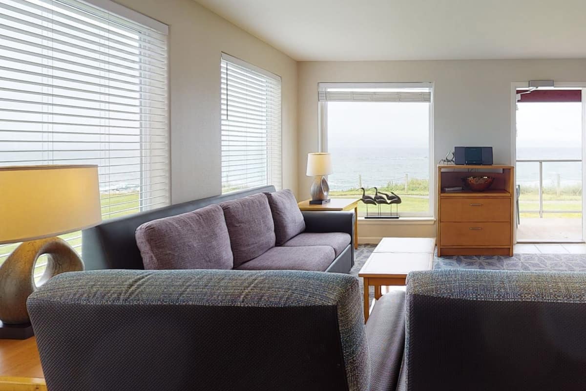 A room with a sofa, a table, and a lamp. There are two windows with blinds, and a view of the outdoors.