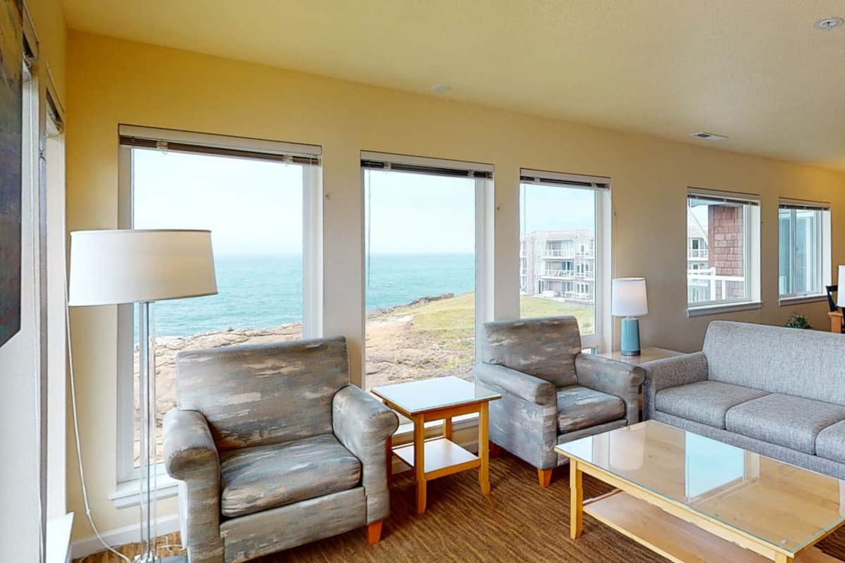 A living room with a view of the ocean, featuring a sofa, armchairs, a coffee table, and a lamp.