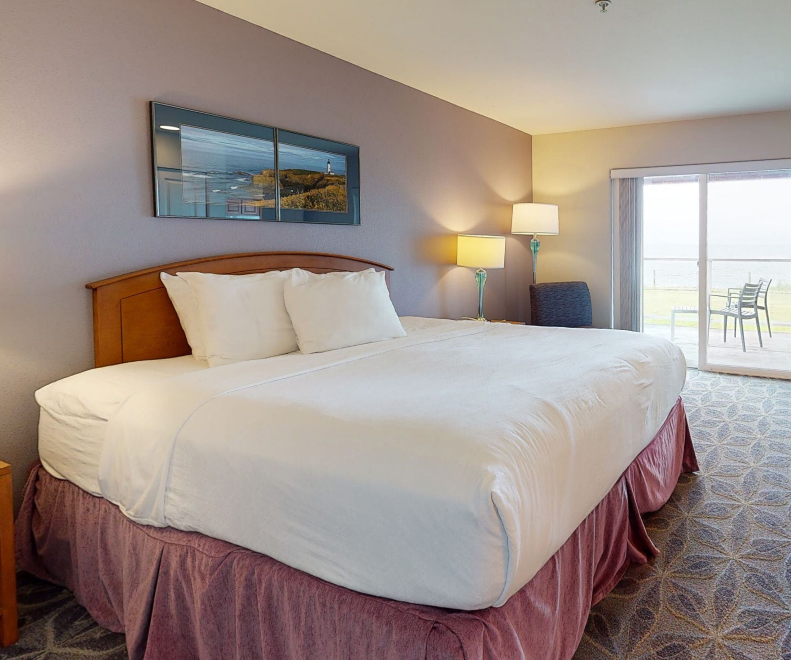 A hotel room with a large bed, a painting on the wall, a lamp, and a view of the outdoors through a sliding door.