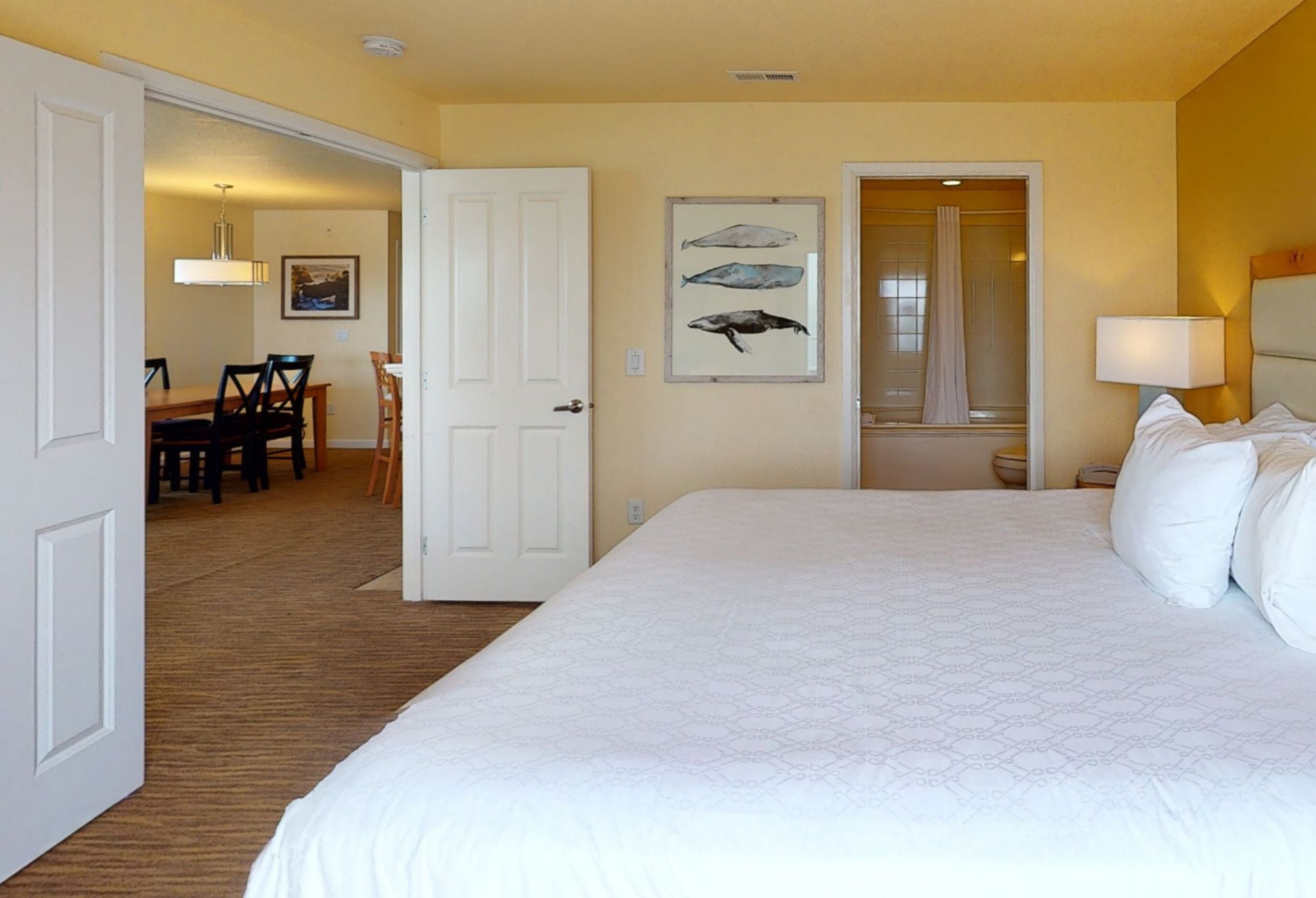 Habitación de hotel con una cama grande, un rincón de comedor con sillas, un cuadro de peces en la pared y un baño visible a través de una puerta abierta.