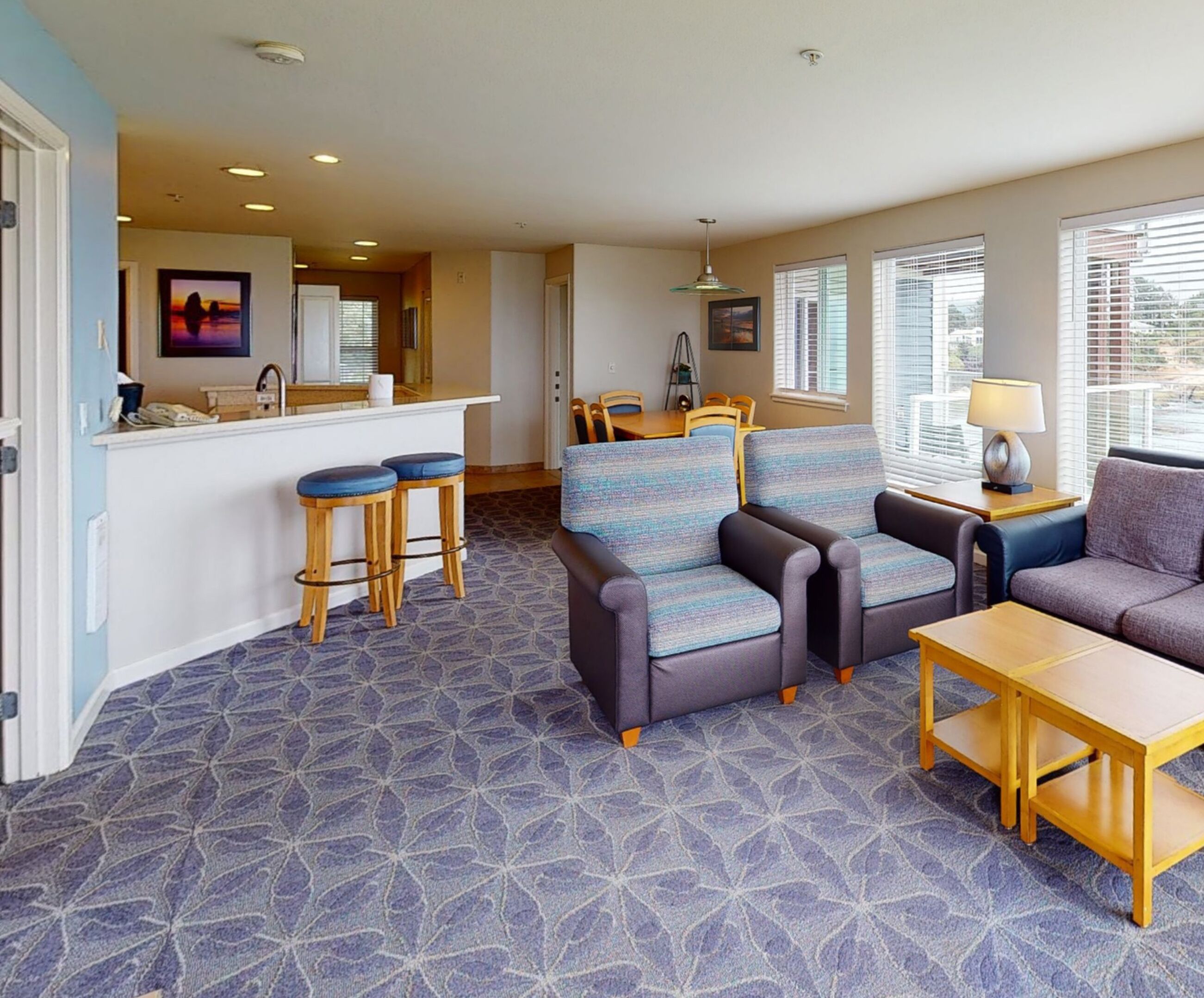 A hotel room with a kitchenette, a seating area with a sofa and chairs, a small table, and a patterned carpet.
