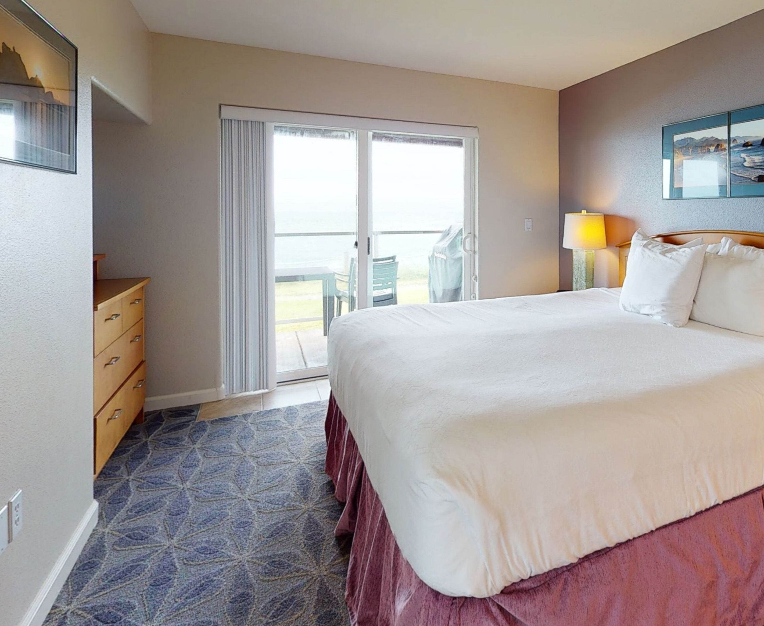 A hotel room with a large bed, a sliding glass door to the balcony, a wooden dresser, and a lamp on the nightstand.
