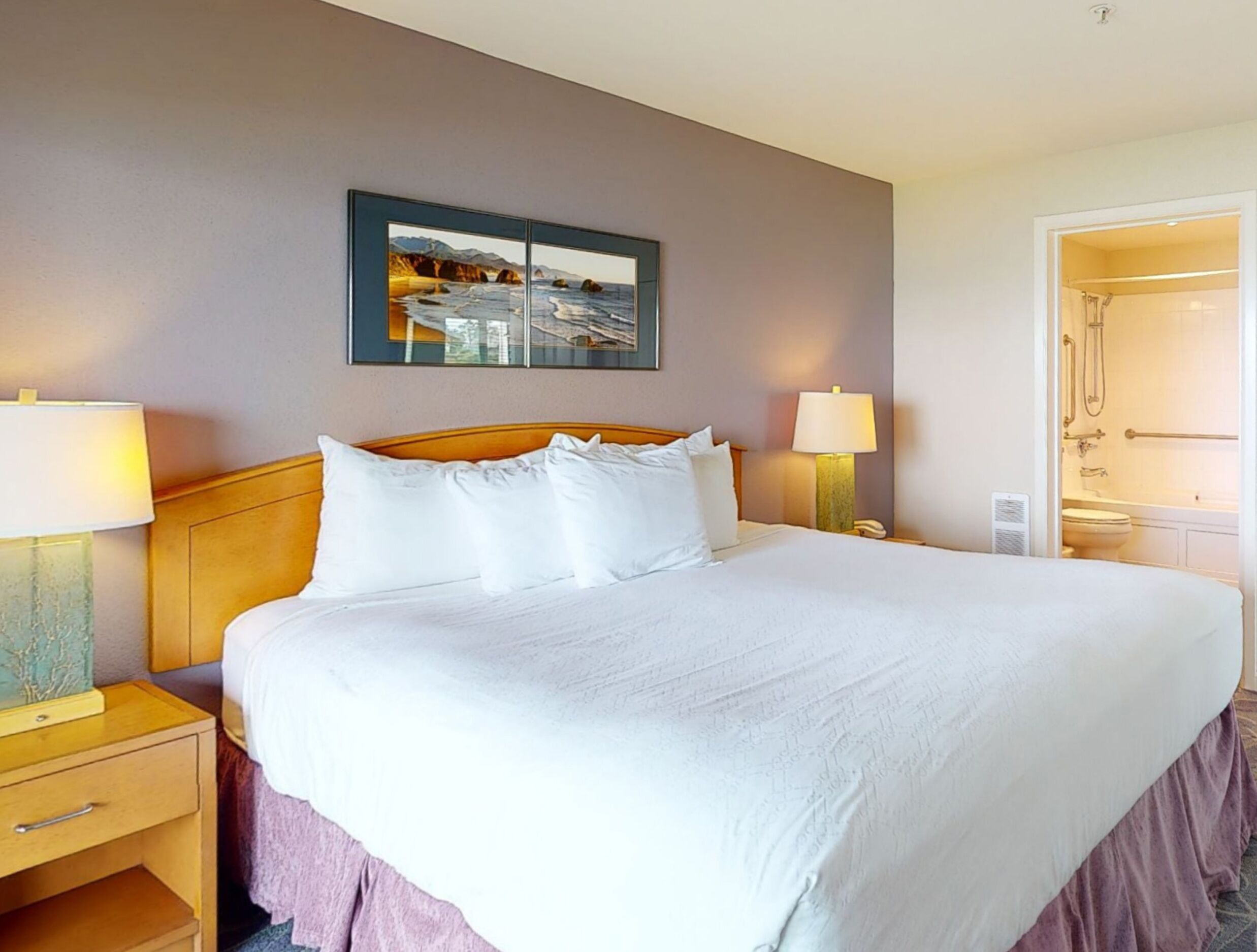 A hotel room with a large bed, two bedside tables with lamps, a nightstand, a painting on the wall, and a bathroom door visible.