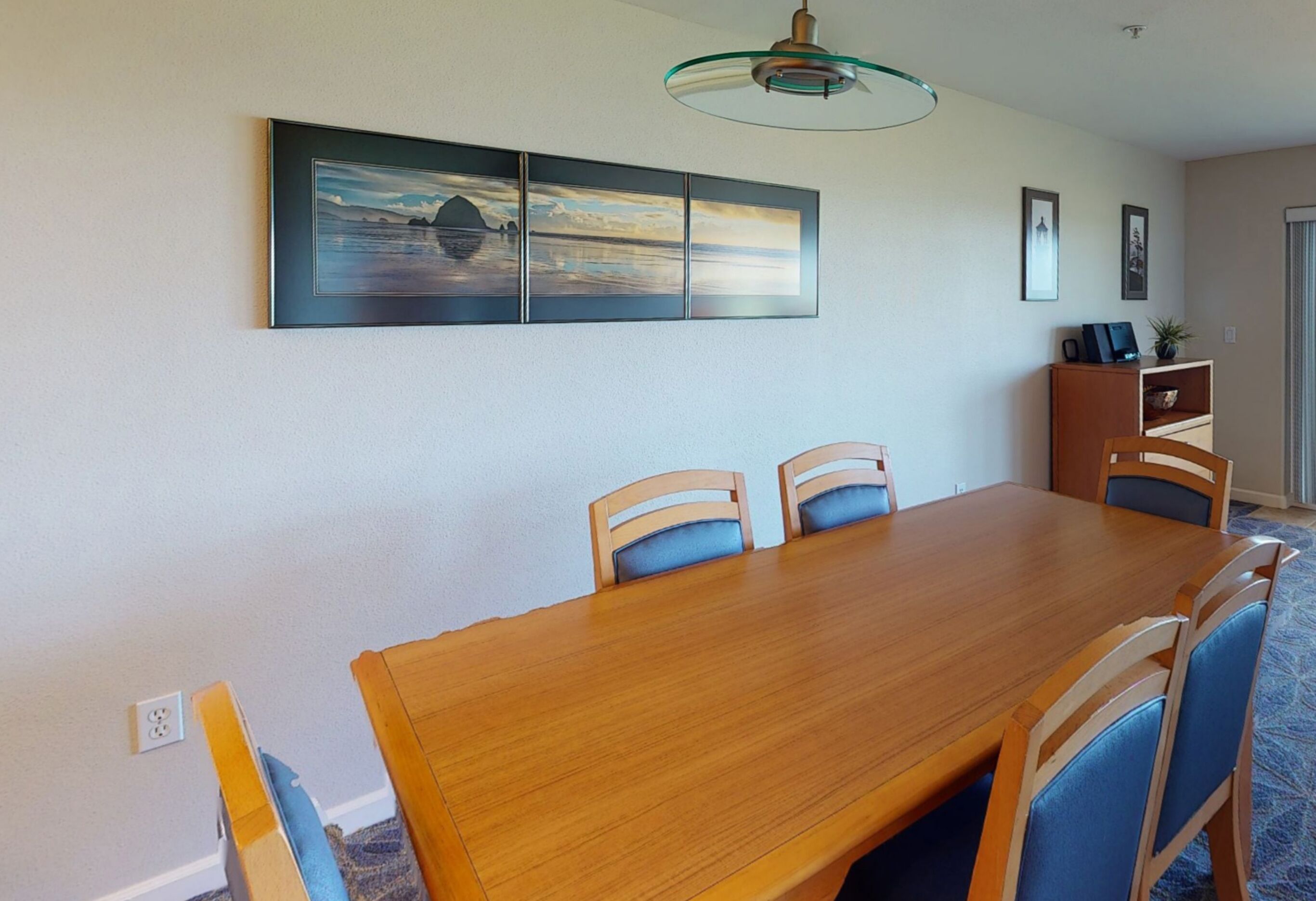 A dining area with a wooden table and blue chairs, a glass ceiling light, and a wall-mounted painting.