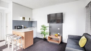 A modern living space with a kitchen area, a grey sofa with a yellow pillow, and a flat-screen TV mounted on the wall. - Metro Hotel Miranda (Miranda)