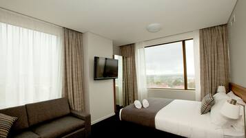 Habitación de hotel con cama, televisor, sofá y amplias ventanas con vistas a la ciudad.