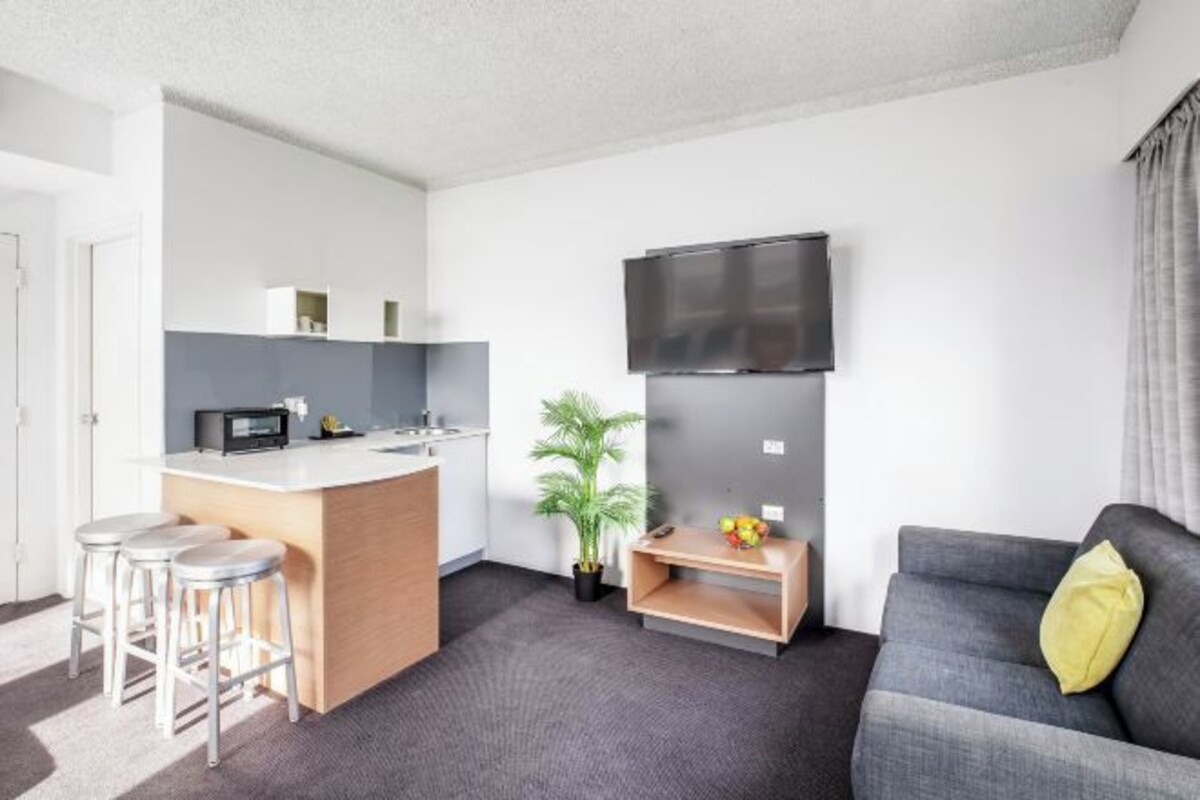 A modern living room with a grey sofa, a small kitchen area with a microwave, and a TV mounted on the wall.