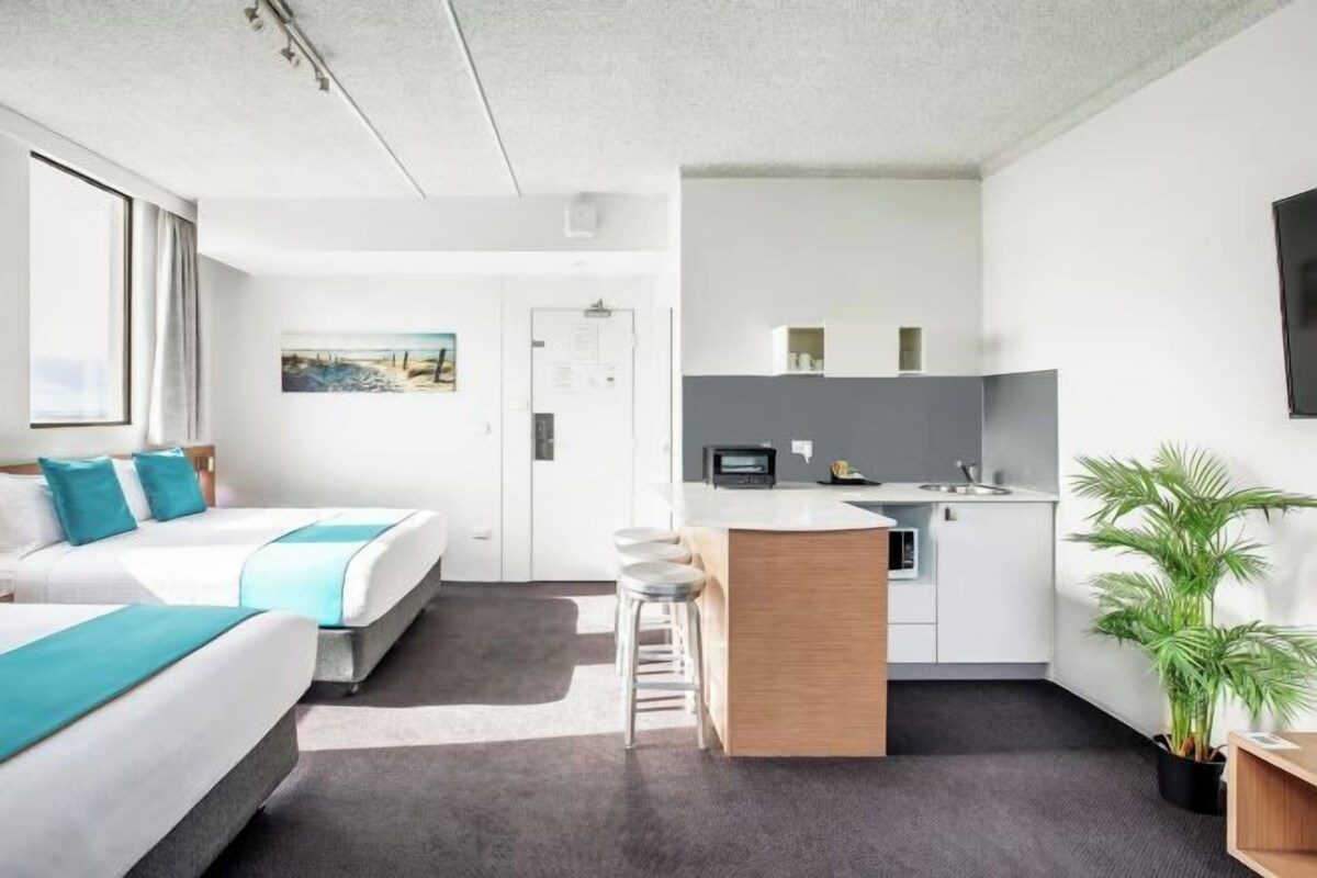 Una habitación de hotel moderna con dos camas, una kitchenette, un pequeño bar y una planta en maceta.