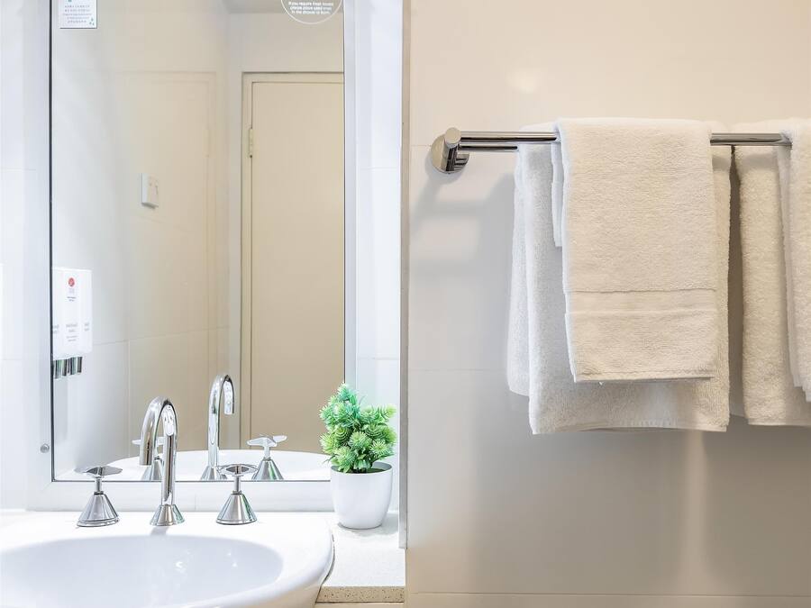 Un baño con un lavamanos blanco, un espejo y un perchero con tres toallas blancas.