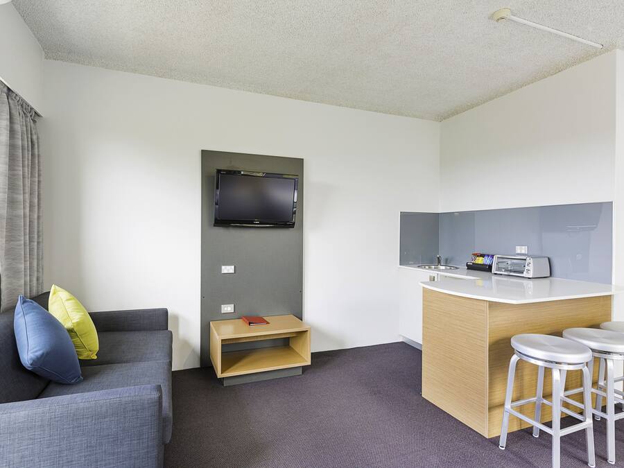 Habitación de hotel compacta con sofá gris, una pequeña mesita de madera, un televisor montado en la pared, una kitchenette con microondas y una pequeña zona de bar con taburetes.