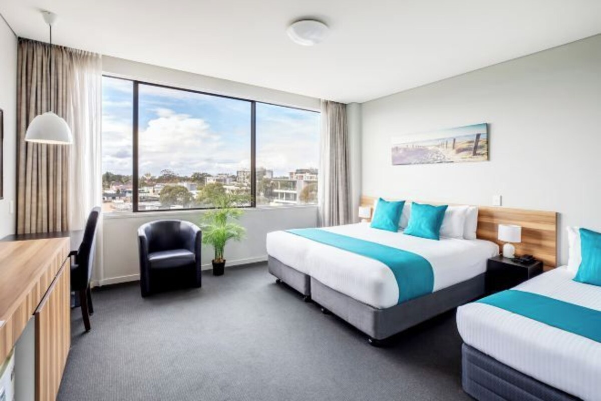 A hotel room with two beds, a desk, a chair, a potted plant, and a large window offering a city view.