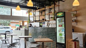 An indoor seating area with tables and chairs, a refrigerated display case, and hanging pendant lights. - Metro Hotel Miranda (Miranda)