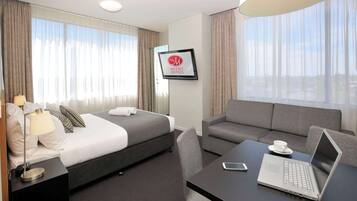 Een hotelkamer met een bed, een bank, een bureau met een laptop, een lamp en een lijst met een kadertje aan de muur.