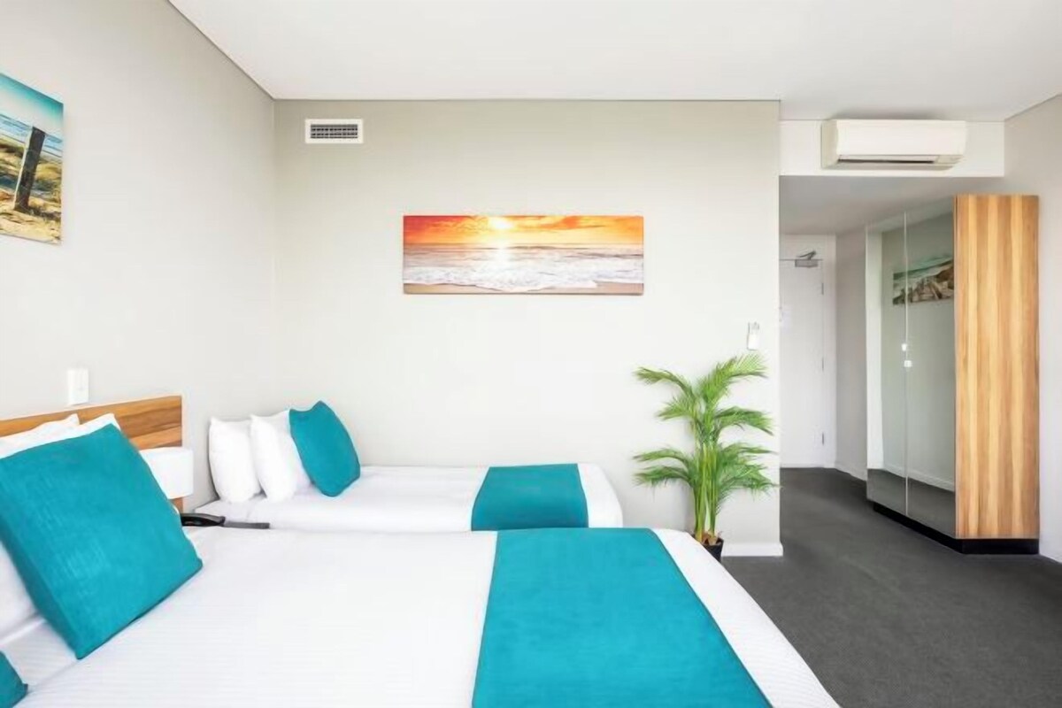 Habitación de hotel con dos camas, un cuadro grande de una puesta de sol, una planta en maceta y un armario.