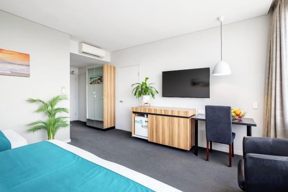 Habitación de hotel con cama, escritorio, silla, televisor y una pequeña kitchenette.