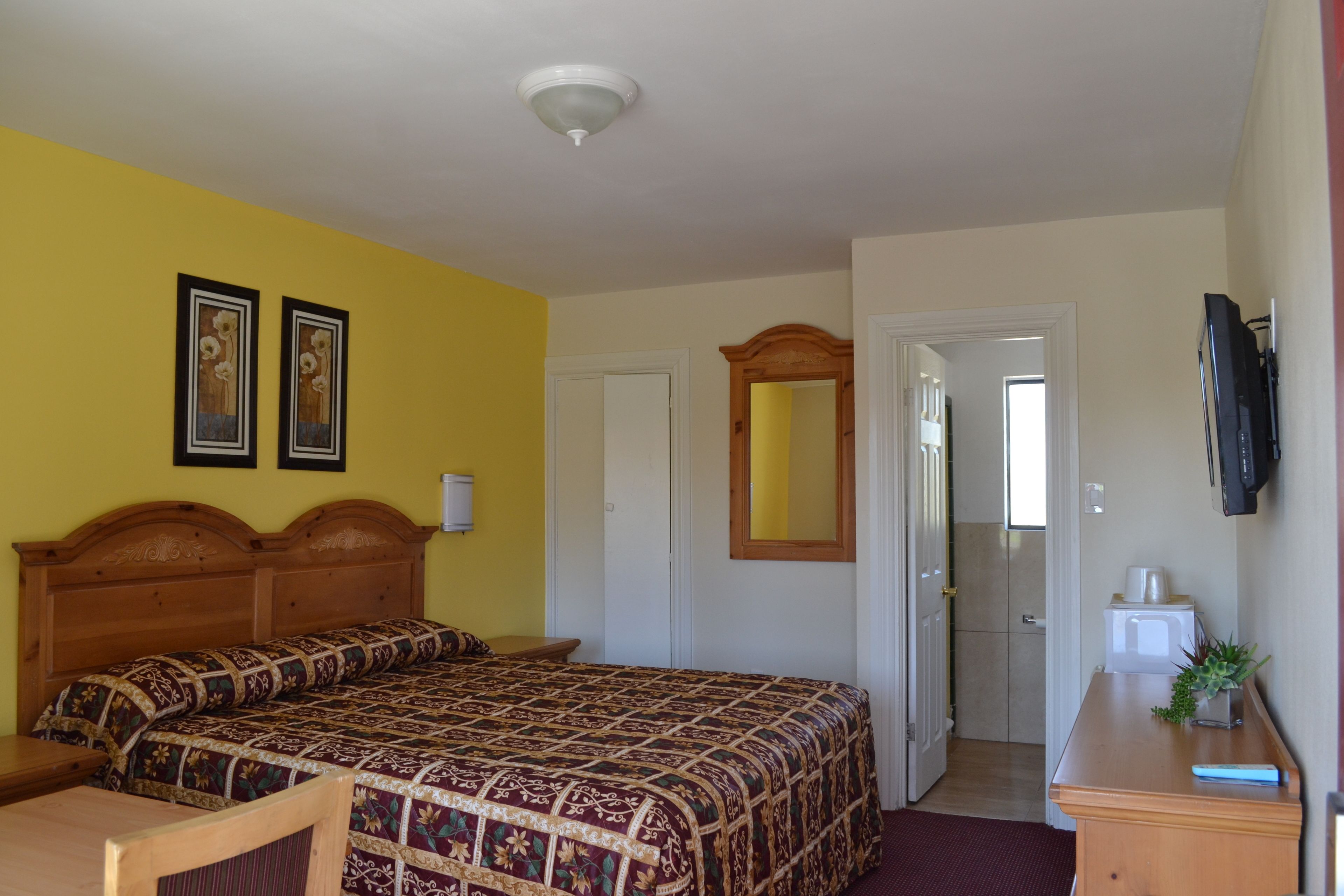 Habitación de hotel con cama de madera, un escritorio con planta, televisor y puerta que da a otra habitación.