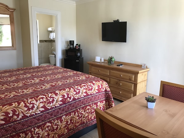 A hotel room with a large bed, a wooden dresser, a television mounted on the wall, and a small table with a plant.