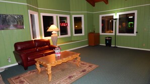 A living room with a brown leather sofa, a wooden coffee table with a vase of flowers, and a lamp. There are two windows with white frames, a door, and a trash can. - Rancho California Inn (Temecula)
