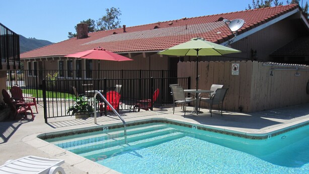 A pool area with a metal ladder, red and green umbrellas, and outdoor furniture. - Rancho California Inn (Temecula)