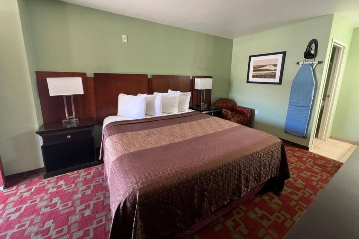 Quarto de hotel com cama grande, mesa de cabeceira, luminária, quadro na parede e um centro de passar roupa azul.