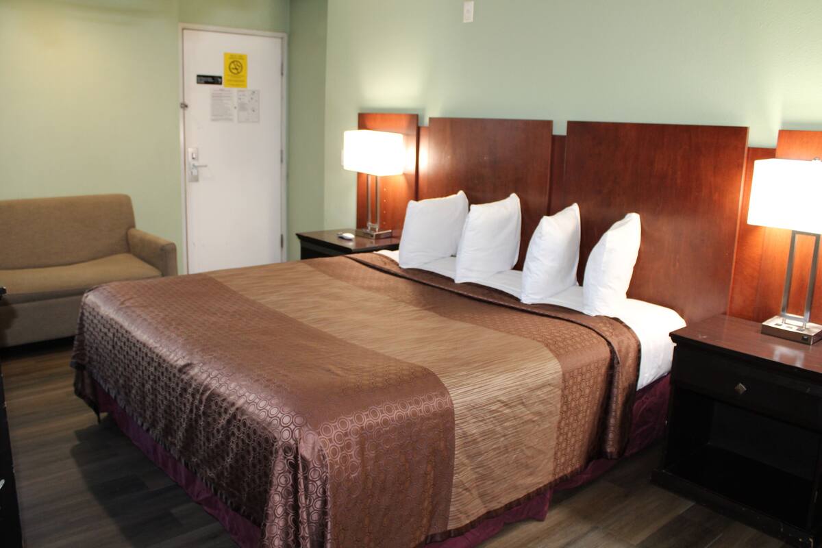 Quarto de hotel com uma cama grande, mesa de cabeceira com abajur, cadeira e porta com placa.