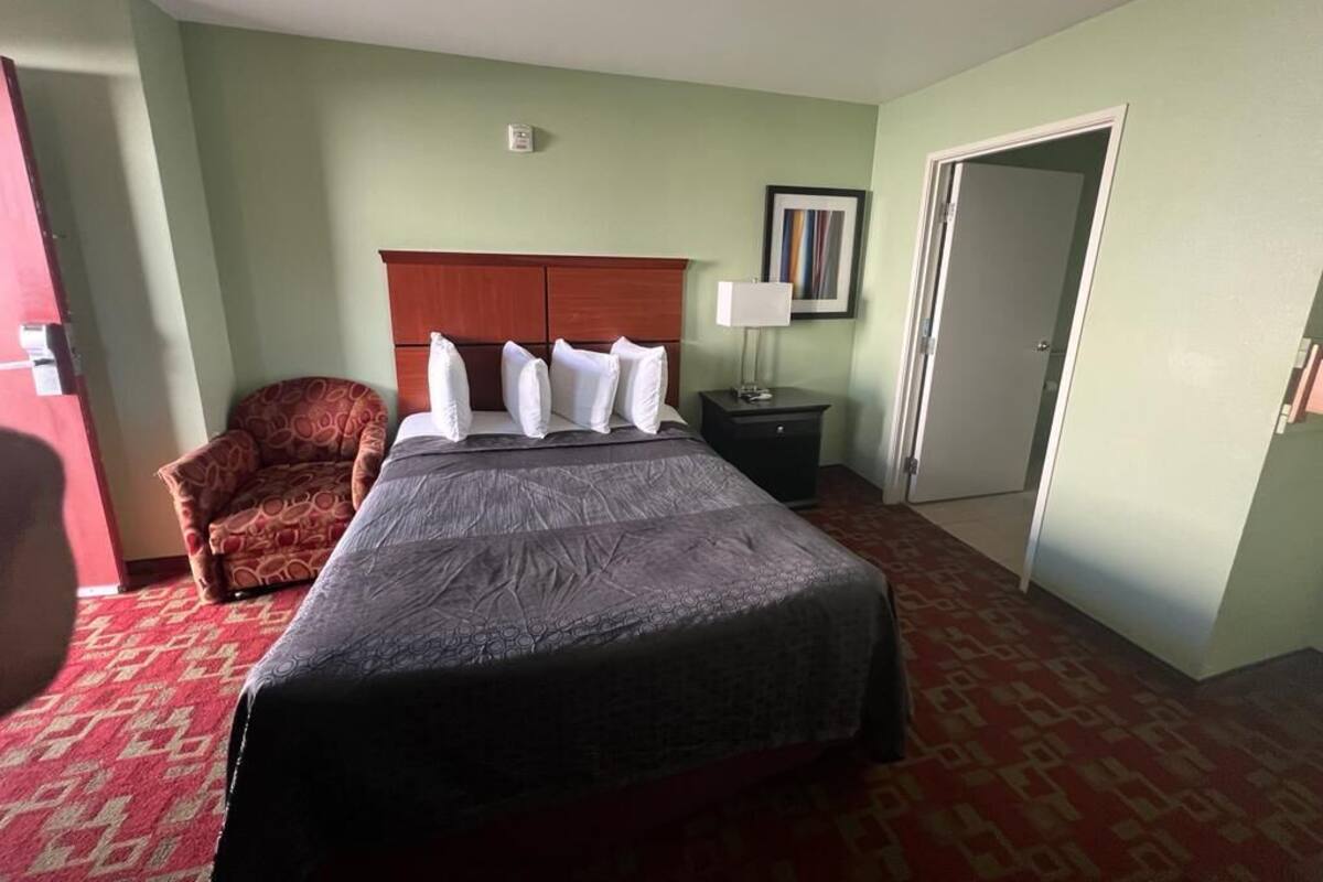 Quarto de hotel com uma cama grande, cabeceira de madeira, carpete vermelho estampado, uma poltrona pequena, mesa de cabeceira com abajur e uma porta.