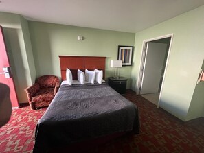 A hotel room with a large bed, a wooden headboard, a red patterned carpet, a small armchair, a nightstand with a lamp, and a door.