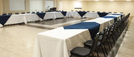 Un salón de conferencias con varias mesas y sillas, manteles blancos y una pantalla para proyector en la pared.