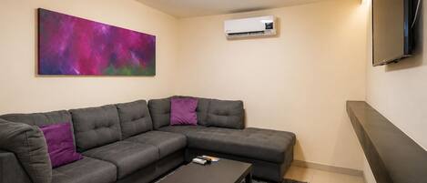 A modern living room with a sectional sofa, a coffee table, a mounted air conditioner, and a flat-screen TV on the wall.
